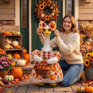 Puede incluir: Una estatua de ganso blanco con un vestido estampado de calabazas y hojas, un lazo a juego y gafas de sol amarillas. El ganso sostiene una pequeña cesta de mimbre llena de calabazas naranjas en miniatura sobre un porche de piedra.