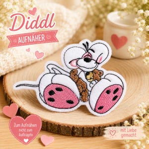 Könnte beinhalten: Ein weißer Stickaufnäher mit der Diddl-Maus-Figur, die große Ohren, rosa Fußsohlen und einen kleinen braunen Teddybären hat. Der Aufnäher zeigt schwarze Konturen und den Text Diddl AUFNÄHER, Zum Aufnähen nicht zum Aufbügeln sowie mit Liebe gemacht.
