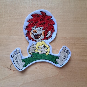 Peut inclure: Écusson brodé représentant un personnage de dessin animé aux cheveux roux hérissés, vêtu d'un haut jaune et d'un pantalon vert. Le personnage sourit, les bras et les jambes écartés. Le patch a une bordure blanche et est destiné aux vêtements ou accessoires.