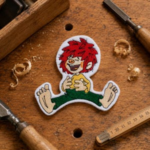 Op de afbeelding: Een geborduurde patch met een stripfiguur met felrood haar, een geel shirt en een groene broek. Het figuur zit op de grond met blote voeten en lacht met de handen op de buik. De patch is voorzien van een witte rand.