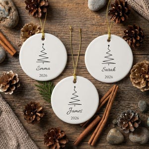 Puede incluir: Tres adornos redondos de cerámica blanca con cordones dorados para colgar. Cada adorno presenta un dibujo lineal negro de un árbol de Navidad, un nombre en letra cursiva y el año 2026 impreso debajo. Los nombres son Emma, Sarah y James.