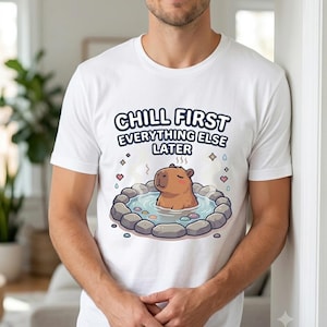 Puede incluir: Una camiseta blanca de manga corta con un gráfico de un capibara marrón relajándose en una fuente termal de piedra. El texto sobre la ilustración dice CHILL FIRST EVERYTHING ELSE LATER en letras azul oscuro, rodeado de pequeños corazones.