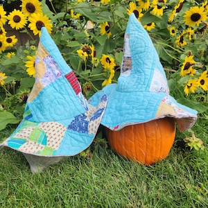 May include: Two pointed hats made from quilted fabric in shades of light blue, tan, and white with various patterns. One hat rests on the ground, while the other is placed on top of a round orange pumpkin, featuring visible stitching.