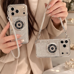 May include: Silver phone cases designed to resemble cameras, embellished with sparkling rhinestones. One case has a wrist strap, the other a shoulder strap. The camera lens details are visible on both cases. The cases are a stylish accessory.