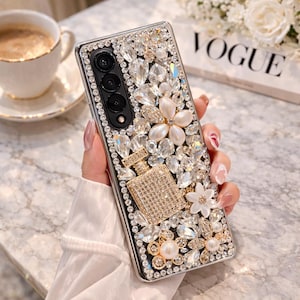 May include: A bedazzled phone case with a black frame, adorned with clear and champagne-colored crystals, pearls, and floral accents. A small, sparkling perfume bottle design is featured on the case. The case is held in a person's hand.