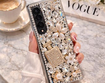 May include: A bedazzled phone case with a black frame, adorned with clear and champagne-colored crystals, pearls, and floral accents. A small, sparkling perfume bottle design is featured on the case. The case is held in a person's hand.