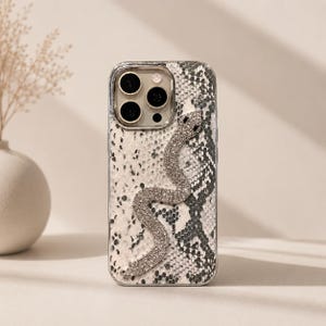 May include: A smartphone case featuring a textured snakeskin pattern in shades of white, gray, and black. A prominent, winding snake-shaped embellishment covered in clear, sparkling rhinestones is centered on the back of the protective cover.