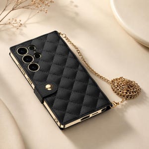 Puede incluir: Una funda de teléfono negra acolchada con una correa de cadena de metal dorado. La funda presenta un patrón de costuras en forma de diamante, un cierre de botón a presión dorado en el lateral y bordes dorados. Las aberturas de la cámara son visibles.