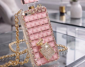 Puede incluir: Funda de teléfono rosa con diseño de frasco de perfume, adornada con pedrería y una correa de cadena dorada. La funda presenta una superficie rosa texturizada y un pequeño dije decorativo de frasco de perfume.