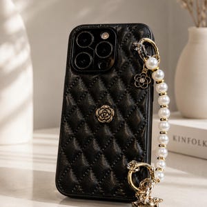 Könnte beinhalten: Eine schwarze, gesteppte Handyhülle mit texturiertem Rautenmuster und einem mittigen, goldfarbenen Rosenemblem. Ein abnehmbares Armband aus weißen Kunstperlen ist befestigt, verziert mit einem kleinen goldenen Blumenanhänger und einem quadratischen Anhänger mit der Zahl 5.