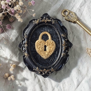 Può includere: Un lucchetto dorato a forma di cuore con dettagli ornati è posto in una cornice ovale nera con accenti dorati. Una chiave dorata è posizionata accanto. L'immagine evoca un tema vintage.
