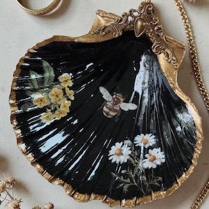 Peut inclure: Un coquillage Saint-Jacques décoratif peint en noir brillant, orné d'un bourdon central, de fleurs jaunes et de marguerites blanches. Les bords de la coquille sont dorés et elle comporte un ornement en métal doré sur la partie supérieure.