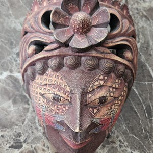 Op de afbeelding: Een gedetailleerd, handgesneden houten masker met een bloemenkroon en ingewikkelde gezichtsdetails. Het masker heeft een donkerbruine basiskleur met lichtere accenten, waaronder een bloem op het voorhoofd en libellenontwerpen op de wangen. Het masker is een decoratief kunstwerk.