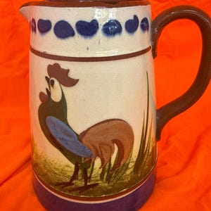 May include: A ceramic pitcher with a rooster design. The pitcher has a white background with a blue and brown rooster illustration. The handle is brown, and the base is blue. The top rim features blue teardrop shapes.
