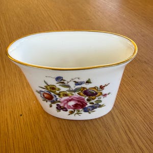 May include: Oval-shaped white ceramic planter with a gold rim and floral pattern. The planter displays a colourful arrangement of pink, blue, yellow, and purple flowers. It is set on a wooden surface.