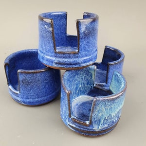 Peut inclure: Trois supports de tasse en céramique bleue avec un design unique. Les supports ont une lèvre surélevée et une découpe au centre. Les supports sont empilés les uns sur les autres.