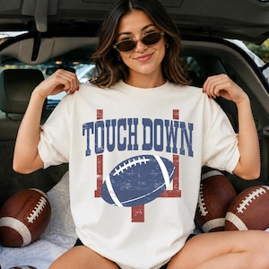 Könnte beinhalten: Ein cremefarbenes Kurzarm-T-Shirt mit einer verwaschenen blauen Grafik eines Footballs und eines Torpfostens sowie dem Schriftzug TOUCH DOWN in großen blauen Blockbuchstaben auf der Brustpartie des Kleidungsstücks.
