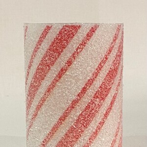 May include: A cylindrical candle holder with a diagonal red and white striped pattern. The surface has a textured, glittery appearance. The design resembles a candy cane, evoking a festive holiday theme.