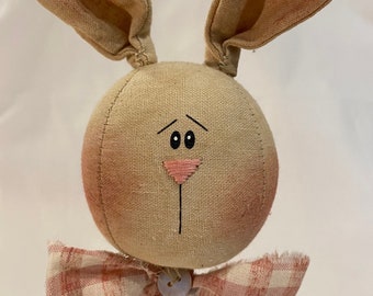 May include: A fabric Easter bunny decoration with long, floppy ears and a round head. The bunny has a pink nose, a black stitched mouth, and a plaid bow. The bunny is made of tan fabric and has a distressed look.