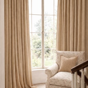May include: Beige linen curtains frame a multi-paned window overlooking a lush green landscape. A floral patterned armchair with a matching pillow sits near the window. The room has a warm, inviting atmosphere.