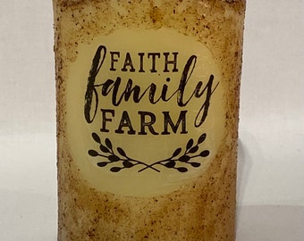 May include: A beige pillar candle with a rustic brown speckled finish. The candle has a white circle label with the text "FAITH family FARM" in black lettering and a pair of crossed sprigs of leaves.