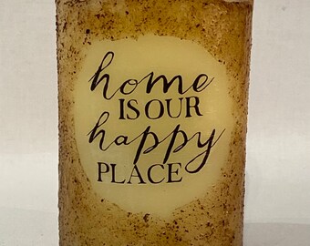 May include: A beige pillar candle with a brown speckled texture. The candle has a white circle with black text that reads "home is our happy place".