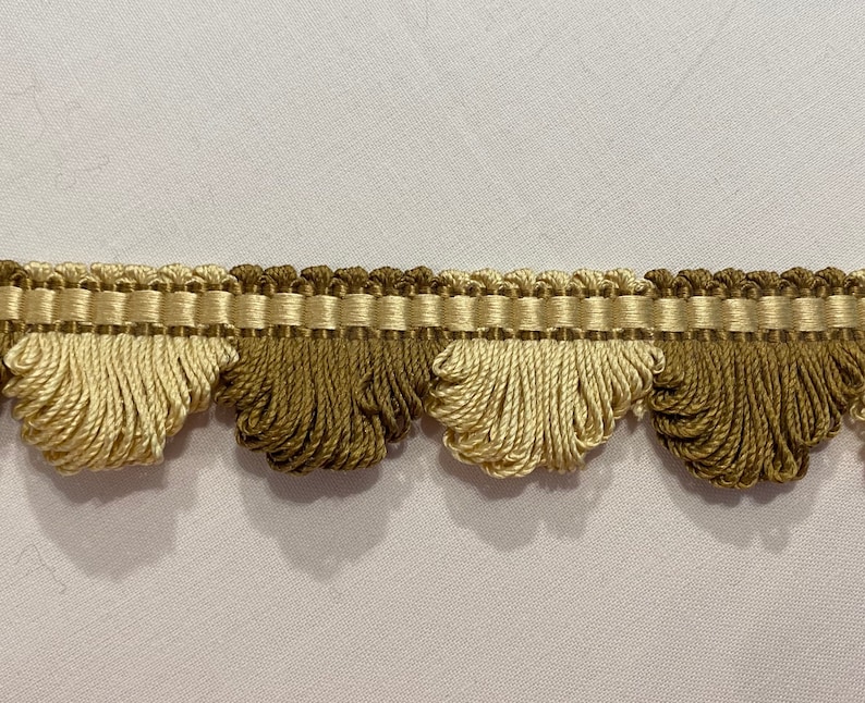 May include: Decorative trim featuring a scalloped design with alternating sections of light and dark beige tassels. The trim has a woven top section in a matching color palette, suitable for home decor and craft projects.