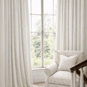May include: A light-filled interior scene featuring floor-to-ceiling, off-white curtains framing a multi-paned window. A floral-patterned armchair with a white pillow sits near the window. The room has a classic, elegant aesthetic.