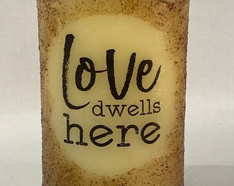 May include: A beige pillar candle with a brown speckled texture and the text "Love dwells here" printed in black.