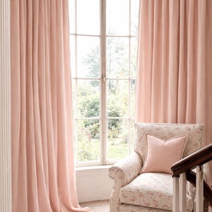 May include: Pale pink curtains frame a large window, with a floral patterned armchair and matching pillow in front. The curtains are floor-length and create a soft, inviting atmosphere. The room has a classic, elegant style.