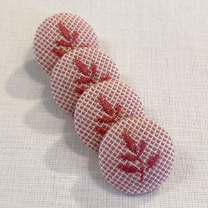 May include: Four pink fabric buttons with a white grid pattern and a red embroidered leaf design.