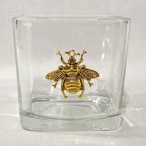 May include: A clear glass square vase with a gold bee design. The bee is detailed with textured wings and a raised body.