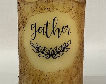 May include: A beige pillar candle with a brown speckled texture. The candle has a white circle with the word "gather" and a black wreath design.