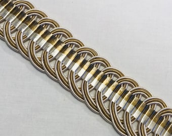 May include: Gold and white braided trim with a looped design. The trim is made of woven fabric and has a subtle sheen.