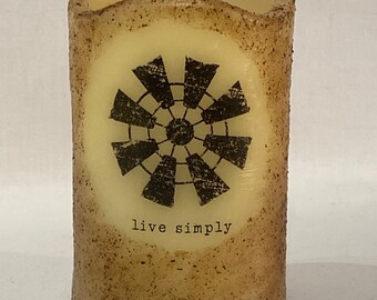 May include: A tan pillar candle with a black and white windmill design and the text "live simply".