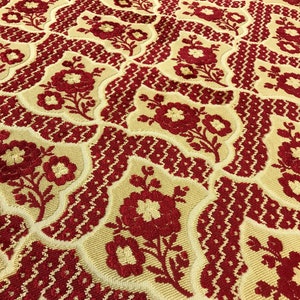 May include: A close-up of a fabric with a repeating pattern of red flowers on a cream background. The fabric has a textured, woven look.
