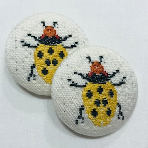 May include: Two white fabric buttons with embroidered yellow and black ladybugs. The ladybugs have red heads and black spots.