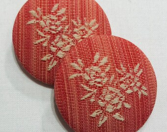 May include: Two red fabric buttons with a white floral design. The buttons are oval shaped and have a textured surface.