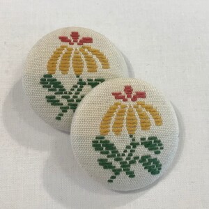 May include: Two round fabric buttons with a yellow flower design with red and green accents. The buttons are white with a subtle texture.