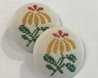 May include: Two round fabric buttons with a yellow flower design with red and green accents. The buttons are white with a subtle texture.