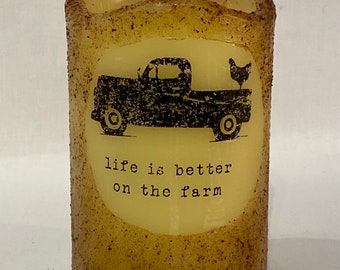 May include: A tan pillar candle with a black and white image of a vintage pickup truck with a chicken in the back. The candle has the text "life is better on the farm" printed on it.