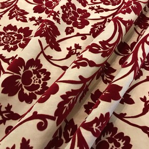 K&ouml;nnte beinhalten: Roter Samtstoff mit wei&szlig;em Hintergrund und einem floralen Muster. Der Stoff hat eine erhabene Textur und ein luxuri&ouml;ses Gef&uuml;hl.