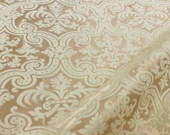 May include: A close-up of a beige fabric with a white damask pattern. The fabric has a subtle sheen and a soft texture.