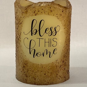 May include: A beige pillar candle with a brown speckled texture. The candle has a white circle with the text "bless this home" in black.