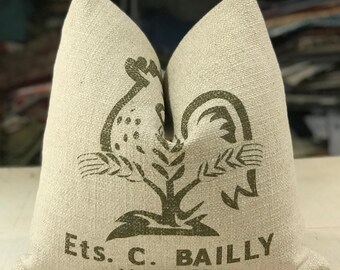 May include: A beige pillow with a green rooster and wheat design. The pillow has the text "Ets. C. BAILLY Marnay 70" printed on it.