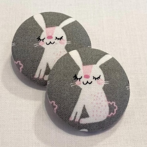 May include: Two gray fabric buttons with white bunnies with pink noses and closed eyes. The bunnies are on a gray background with white accents.