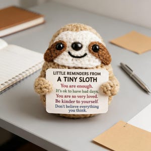 May include: A crocheted sloth figurine with a white face, brown accents, and black eyes, holding a card with the text "LITTLE REMINDERS FROM A TINY SLOTH". The card includes positive affirmations. The sloth is on a gray surface.
