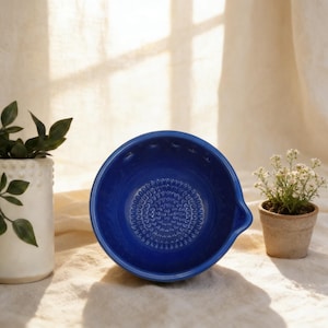 May include: A vibrant cobalt blue ceramic grater bowl with a textured interior. The bowl features a small spout. A white vase with green leaves and a small potted plant with white flowers are also in the image.
