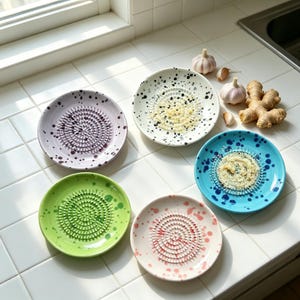 May include: A collection of colorful ceramic garlic graters. Each plate features a central grater and is speckled with paint. Colors include purple, white, blue, green, and pink. Garlic and ginger are also present.