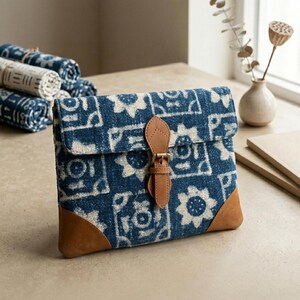 Pode incluir: Uma clutch estampada em azul e branco com fecho de fivela de couro e detalhes em couro castanho. A clutch tem uma aba e é feita de um tecido texturizado. Têxteis enrolados são visíveis no fundo.